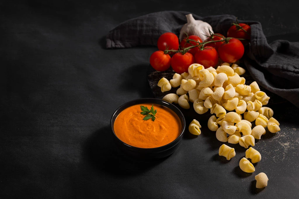 Rosé sauce in a black bowl beside fresh lumaconi and tomatoes